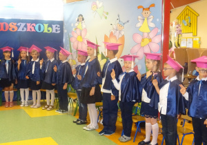 Dzieci stoją w półkolu, podnoszą dwa palce prawej ręki do góry