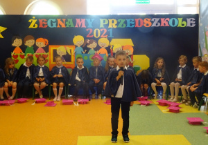 Chłopiec stoi na środku holu, trzyma mikrofon, dzieci siedzą w półkolu