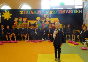 Franek stoi na środku holu, trzyma mikrofon, dzieci siedzą w półkolu