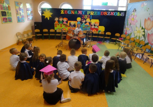 Filip gra na perkusji, dzieci siedzą przed nim w siadzie skrzyżnym
