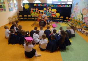 Filip gra na perkusji, dzieci siedzą przed nim w siadzie skrzyżnym