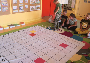 Na dywanie leży mata do kodowania. Dzieci siedzą wokół niej. Po prawej stronie na dywanie rozsypane są kody zapisane na karteczkach. Chłopiec wybiera kod do odszukania.