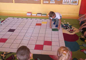 Dzieci siedzą wokół maty ułożonej na dywanie i obserwują kolegę który zaznacza na macie zieloną tabliczką określone pole.