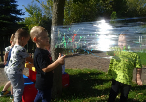 Dzieci malują paluszkami na foli rozłożonej pomiędzy drzewami.