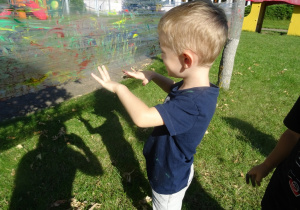 Chłopiec maluje paluszkami na foli rozłożonej pomiędzy drzewami.