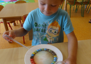 Chłopiec nalewa wodę pipetą na talerz. Obserwuje zjawisko rozpuszczania cukierków