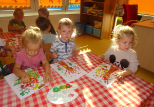 Dzieci stemplują paluszkami maczanymi w farbie wokół odbitej rączki na kartce tworząc jesienne drzewo.