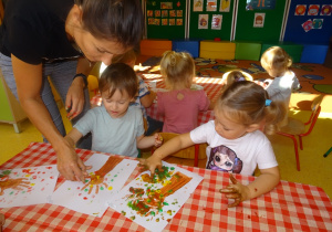 Dzieci stemplują paluszkami maczanymi w farbie wokół odbitej rączki na kartce tworząc jesienne drzewo.
