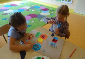 Dwie dziewczynki wykonują pracę plastyczną dowolną techniką, malują farbami, naklejają elementy z papieru.