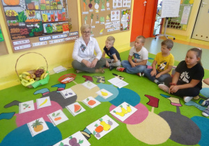 Dzieci rozwiązują zagadki, na dywanie rozłożone są ilustracje owoców.