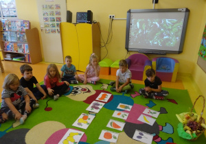Dzieci siedzą wokół ilustracji owoców.