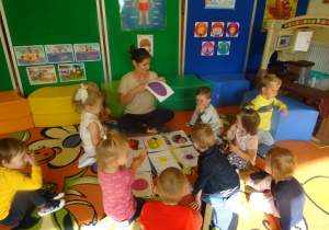 Dzieci siedzą na dywanie wokół pani Eweliny która prezentuje plansze z kształtami.