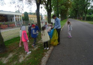 Dziewczynka wrzuca śmieci do worka, który trzyma pani Monika. W tle dzieci zbierają śmieci z trawnika.