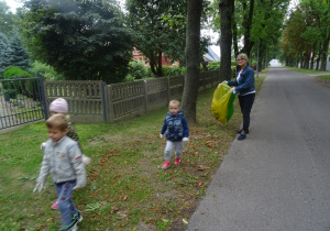 Troje dzieci spaceruje po trawniku wraz z panią Basią, która trzyma worki na śmieci.