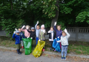 Dziewięcioro dzieci pozuje do zdjęcia z uniesionymi rękami na których mają założone rękawice. Troje dzieci trzyma worki z zebranymi śmieciami.