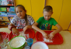 Dzieci wykonują doświadczenie z cieczą nienewtonowską, mieszają mąkę z wodą na tackach.