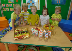 Uśmiechnięte dzieci stoją wokół stolika z owocowymi przekąskami.