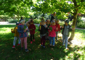 Grupa dzieci stoi z liśćmi uniesionymi w dłoniach. W tle drzewa.
