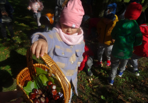Dziewczyna trzyma w ręku koszyk z darami jesieni. W tle dzieci zbierające liście.