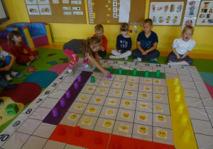 Dzieci grają w grę "Emocje" na macie edukacyjnej, dziewczynka przestawia kubek na wybrane pole, reszta dzieci siedzi wokół maty i przygląda się.