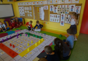 Dzieci grają w grę "Emocje" na macie edukacyjnej, dziewczynka rzuca kostką, reszta dzieci siedzi wokół maty.