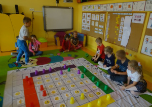 Dzieci grają w grę "Emocje" na macie edukacyjnej, chłopiec rzuca kostką, reszta dzieci siedzi wokół maty.