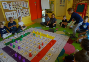 Dzieci grają w grę "Emocje" na macie edukacyjnej, chłopiec rzuca kostką, reszta dzieci siedzi wokół maty.