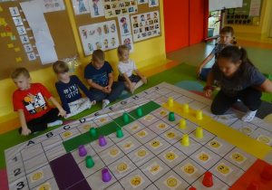 Dzieci grają w grę "Emocje" na macie edukacyjnej, dziewczynka przekłada kubek w określone miejsce, reszta dzieci siedzi wokół chusty.