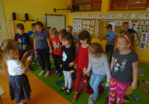 Dzieci stoją na przed dziewczynką która prezentuje obrazek z wybraną emocją, którą dzieci pokazują za pomocą ruchu, gestu, mimiki twarzy.