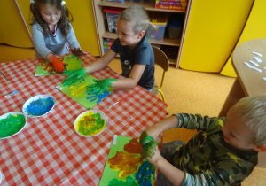 Dwójka dzieci odbijają rączki na kartce, jedne chłopiec pociera swoje rączki wymalowane farbą.