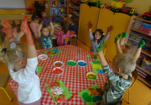 Szóstka dzieci siedzi przy stoliku z uniesionymi rączki pomalowanymi farbą.
