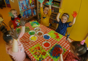 Piątka dzieci siedzi przy stoliku z uniesionymi rączki pomalowanymi farbą.