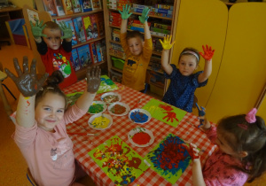 Piątka dzieci siedzi przy stoliku z uniesionymi rączki pomalowanymi farbą.