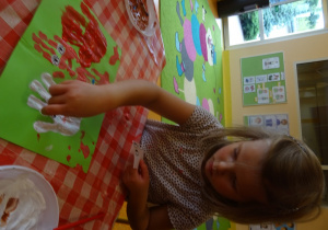 Dziewczynka dokleja oczy do odbitych na kartce papieru dłoni.