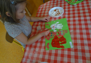 Dziewczynka domalowuje pędzlem czerwone usta do odbitych na kartce papieru dłoni.