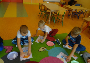 Trójka dzieci siedzi na dywanie, każdy układa obrazek z części.