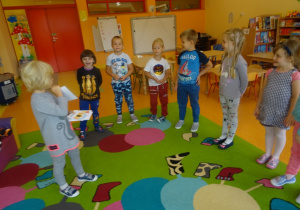 Dzieci stoją w półkolu , dziewczynka w środku pokazuje emocje na obrazku a reszta dzieci pokazują ją za pomocą mimiki twarzy