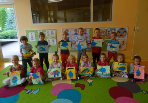 Dzieci stoją lub siedzą na dywanie ze swoimi pracami plastycznymi.