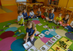Dziewczynka dopasowuje ilustracje drzewa, liść i owoc. W tle przyglądają się jej siedzące na dywanie dzieci.