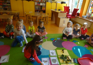 Dwie dziewczynki dopasowują ilustracje drzewa, liść i owoc. Grupa dzieci siedzi wokół na dywanie.