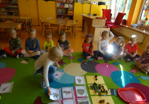 Dziewczynka dopasowuje ilustracje drzewa, liść i owoc. W tle grupa dzieci siedzi na dywanie.