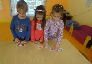 Troje dzieci stoi przy stoliku, dobiera pary pasujących obrazków do siebie darów jesieni.