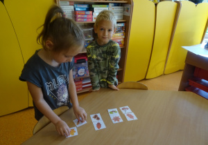 Dwoje dzieci stoi przy stoliku, dobiera pary pasujących obrazków do siebie darów jesieni.