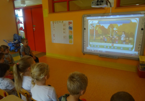 Dzieci siedzą na krzesełkach wokół tablicy interaktywnej, oglądają film edukacyjny