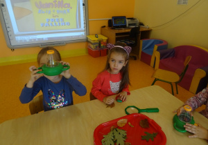 Dwójka dzieci siedzi przy stoliku, oglądają przez lupy liście.