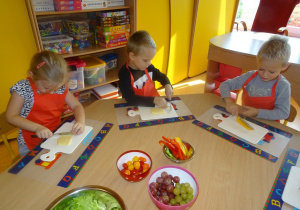 Troje dzieci siedzi przy stoliku w ręku trzymają nóż, którym kroją żółty ser oraz paprykę na deskach.