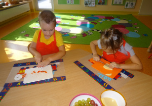 Dwoje dzieci siedzi przy stole, w prawej ręce trzymają nóż którym kroją żółty ser i paprykę na deskach.