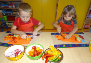 Dwoje dzieci siedzi przy stole, w ręku trzymają nóż którym kroją paprykę na deskach. Na środku stołu stoją miski wypełnione warzywami przygotowanymi do krojenia przez dzieci.