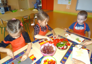 Troje dzieci siedzi przy stole, w ręku trzymają nóż którym kroją warzywa. Na środku stołu stoją miski wypełnione winogronem i warzywami przygotowanymi do krojenia.