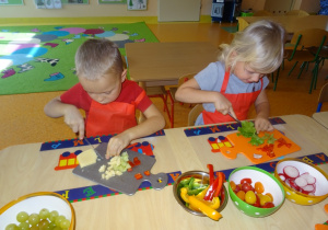 Dwoje dzieci siedzi przy stole, w ręku trzymają nóż którym kroją sałatę i żółty ser.
